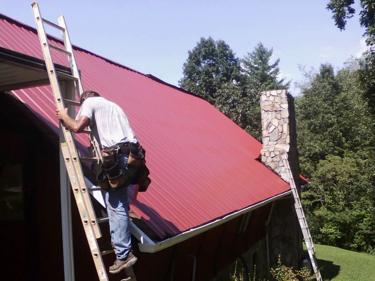 Roofer on a ladder beside a red metal roof and stone chimney — Soffit & Fascia Repair by Roofing Brothers in Winnett